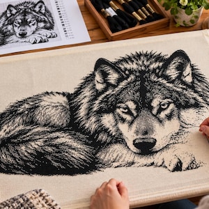 Op de afbeelding: Een kruissteek borduurproject op lichtbeige stof met een gedetailleerde zwart-witillustratie van een rustende wolf. Het ontwerp toont de wolf liggend met de kop naar voren, uitgevoerd in gedetailleerde zwarte draadsteken.