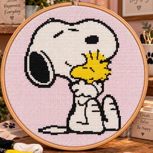 Könnte beinhalten: Ein runder Stickrahmen aus Holz mit einem Kreuzstichmuster, das den weißen Hund Snoopy zeigt, wie er den gelben Vogel Woodstock vor einem hellrosa Hintergrund umarmt. Das Design besteht aus schwarzen Umrissen und vollflächigen Stichen.