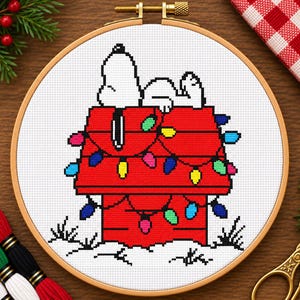 May include: A cross-stitch embroidery in a round wooden hoop featuring a white cartoon dog lying on top of a red doghouse. The house is decorated with a string of colourful Christmas lights in blue, green, yellow, and pink, set against white fabric.