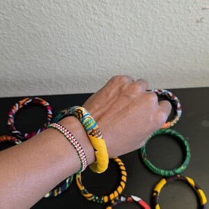 May include: A collection of colorful fabric-covered bangles. One wrist displays a yellow and patterned bangle, and a beaded bracelet. Other bangles with various patterns and colors are scattered on a black surface.