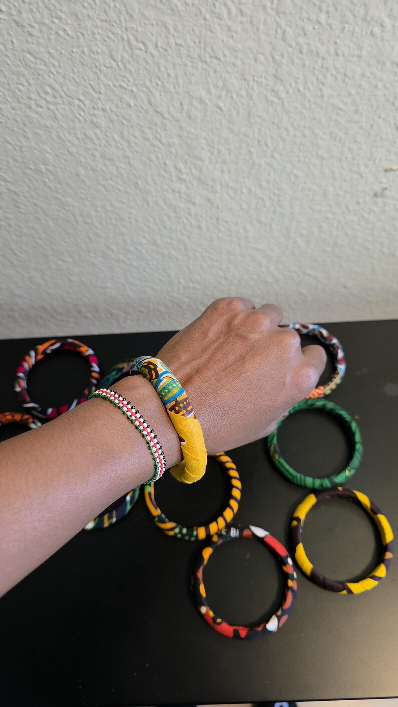 May include: A collection of colorful fabric-covered bangles. One wrist displays a yellow and patterned bangle, and a beaded bracelet. Other bangles with various patterns and colors are scattered on a black surface.