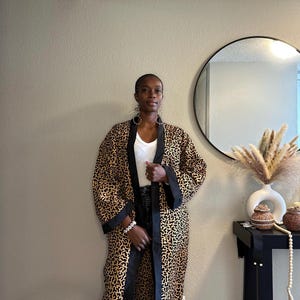 May include: A long, open-front robe with a leopard print pattern in shades of brown and black. The robe has black trim along the edges and sleeves. The person is wearing a white top and dark jeans. A round mirror and decorative items are in the background.