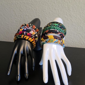May include: A display of fabric-covered headbands in various colorful patterns. The headbands are stacked on black and white mannequin hands. The patterns include vibrant yellows, oranges, blues, and greens.