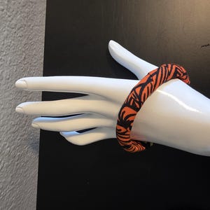 May include: An orange and black patterned fabric bracelet. The bracelet is wrapped around a white mannequin hand. The pattern is a repeating design of black lines on an orange background. The background is black.