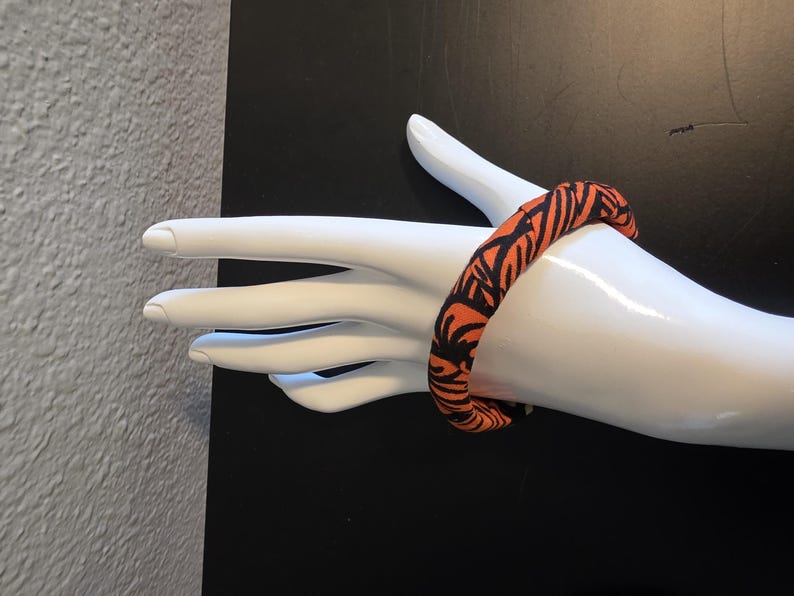 May include: An orange and black patterned fabric bracelet. The bracelet is wrapped around a white mannequin hand. The pattern is a repeating design of black lines on an orange background. The background is black.