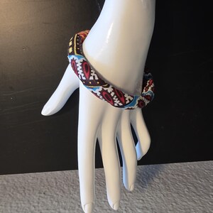 May include: A colorful fabric bracelet with a repeating pattern of red, white, blue, and yellow geometric shapes. The bracelet is displayed on a white mannequin hand against a black background.