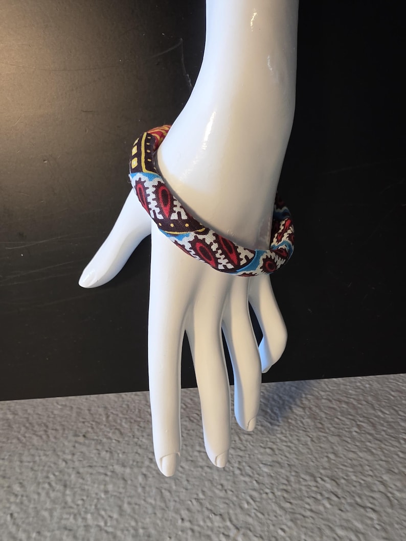 May include: A colorful fabric bracelet with a repeating pattern of red, white, blue, and yellow geometric shapes. The bracelet is displayed on a white mannequin hand against a black background.