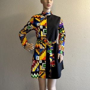 May include: A long-sleeved dress featuring a striking geometric pattern in vibrant colors on one side and solid black on the other. The dress includes a matching belt and a collared neckline. The dress is displayed on a mannequin.