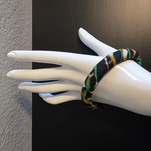 May include: A fabric-covered bangle bracelet in navy blue, green, and gold. The bracelet is displayed on a white mannequin hand against a black background. The bangle has a repeating pattern.