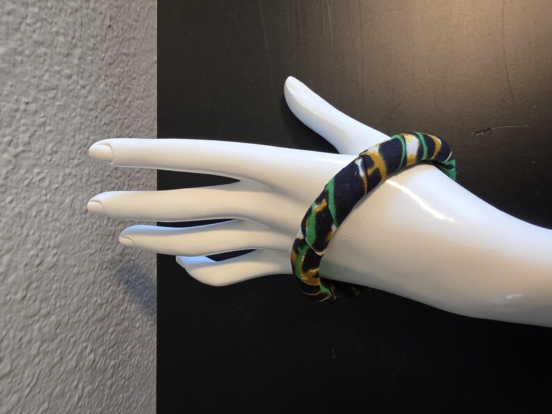 May include: A fabric-covered bangle bracelet in navy blue, green, and gold. The bracelet is displayed on a white mannequin hand against a black background. The bangle has a repeating pattern.