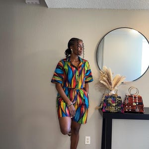 May include: A vibrant two-piece outfit comprising a button-down shirt and matching shorts. The fabric displays a colourful pattern of blue, yellow, and red feather-like designs. Two decorative handbags with gold handles are on a black table.