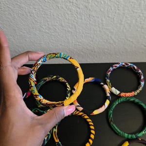 May include: A collection of fabric-covered bangles in various colors and patterns. The bangles are primarily yellow, blue, green, and red, with geometric and abstract designs. One bangle is held by a hand.