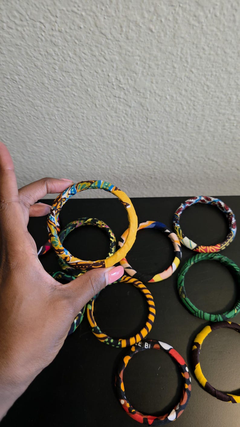 May include: A collection of fabric-covered bangles in various colors and patterns. The bangles are primarily yellow, blue, green, and red, with geometric and abstract designs. One bangle is held by a hand.