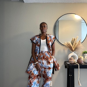 May include: A woman wearing a matching two-piece outfit. The outfit includes a sleeveless jacket and wide-leg pants. The fabric features a brown, blue, and white geometric pattern. The jacket has black trim, and the woman is wearing a white top underneath.