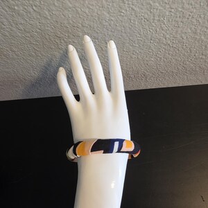 May include: A colorful bangle bracelet with a geometric pattern of navy, yellow, and orange on a white mannequin hand. The bracelet is a thick, rounded design, showcasing a vibrant mix of colors and shapes. The background is a neutral gray and black.