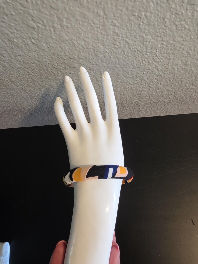 May include: A colorful bangle bracelet with a geometric pattern of navy, yellow, and orange on a white mannequin hand. The bracelet is a thick, rounded design, showcasing a vibrant mix of colors and shapes. The background is a neutral gray and black.