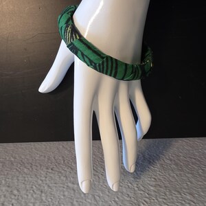 May include: A green and black patterned fabric bracelet. The bracelet is wrapped around a white hand. The fabric has a repeating pattern of black stripes on a green background. The bracelet is a simple, yet stylish accessory.