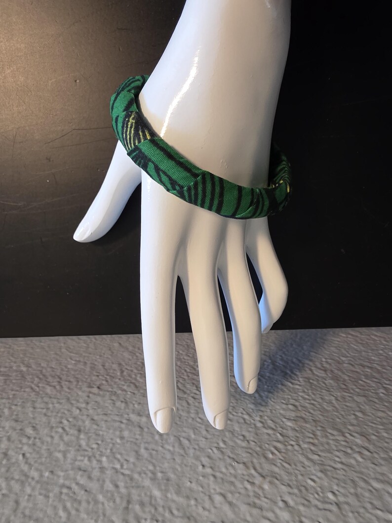 May include: A green and black patterned fabric bracelet. The bracelet is wrapped around a white hand. The fabric has a repeating pattern of black stripes on a green background. The bracelet is a simple, yet stylish accessory.
