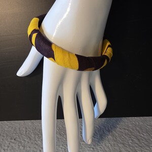 May include: A fabric-covered bangle bracelet in yellow and dark brown. The bracelet is wrapped in a repeating pattern of yellow and dark brown shapes. The bracelet is displayed on a white mannequin hand.