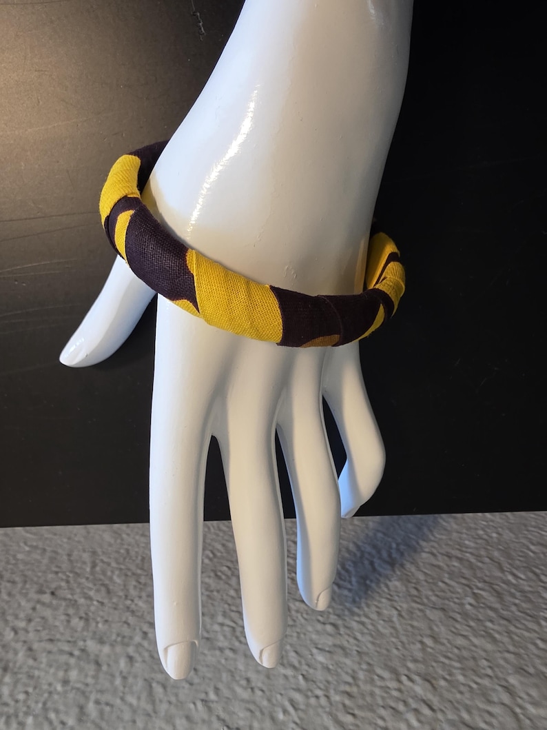 May include: A fabric-covered bangle bracelet in yellow and dark brown. The bracelet is wrapped in a repeating pattern of yellow and dark brown shapes. The bracelet is displayed on a white mannequin hand.