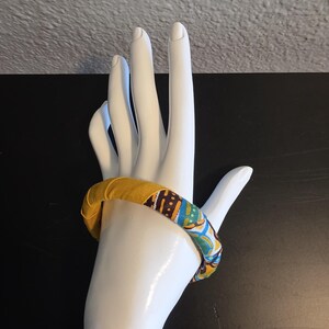 May include: A fabric-covered bangle bracelet in shades of yellow, blue, and brown. The bracelet is displayed on a white hand-shaped mannequin. The fabric has a patterned design.