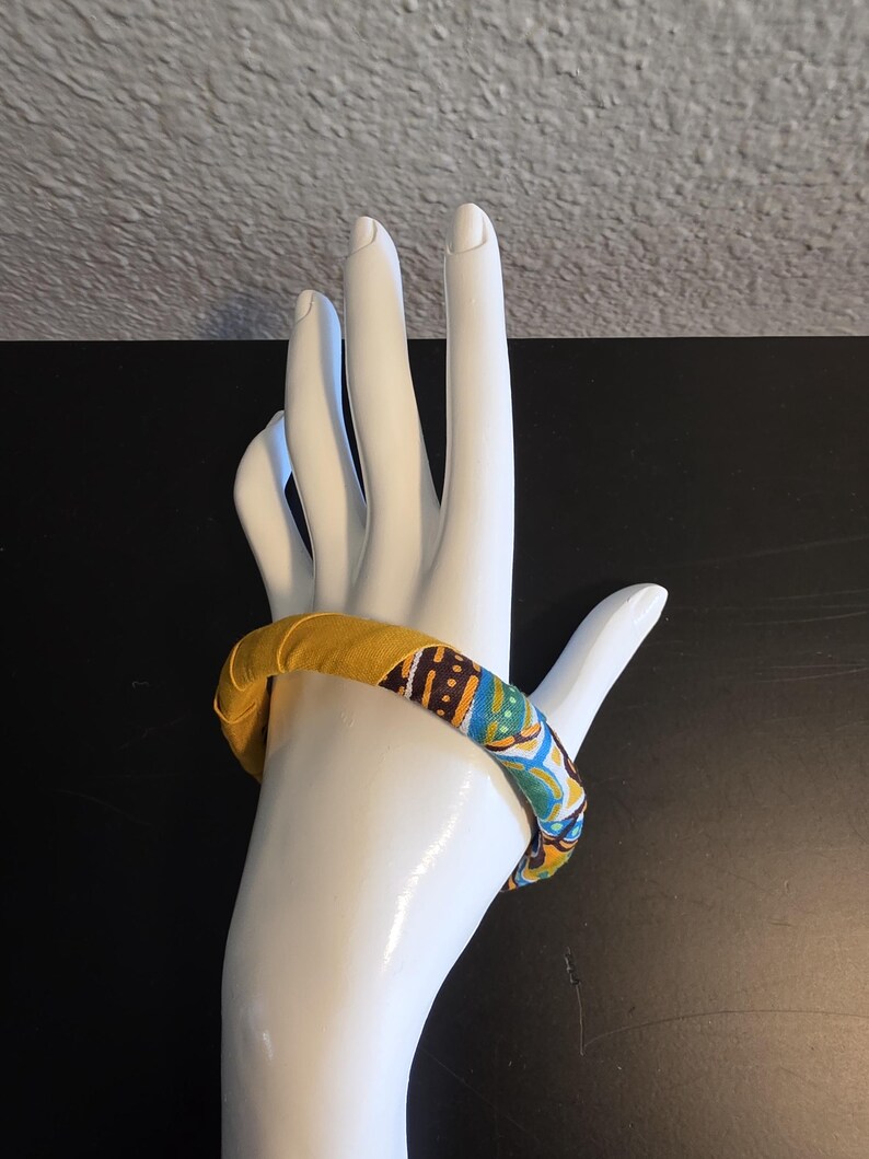 May include: A fabric-covered bangle bracelet in shades of yellow, blue, and brown. The bracelet is displayed on a white hand-shaped mannequin. The fabric has a patterned design.