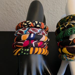 May include: A stack of six fabric-covered bangles with vibrant patterns in orange, black, red, blue, and yellow. The bracelets are displayed on a black hand-shaped jewelry holder, showcasing the colorful designs.