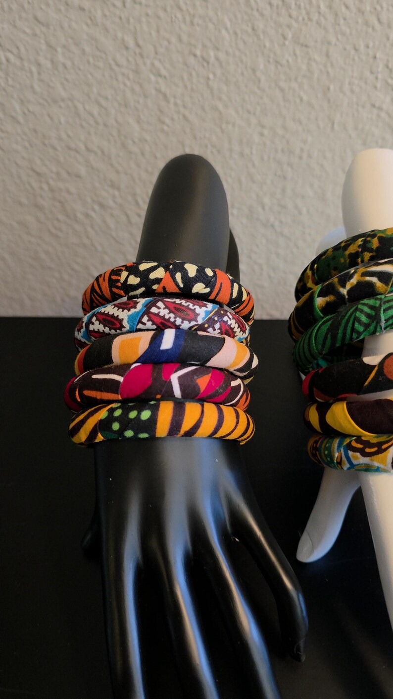 May include: A stack of six fabric-covered bangles with vibrant patterns in orange, black, red, blue, and yellow. The bracelets are displayed on a black hand-shaped jewelry holder, showcasing the colorful designs.