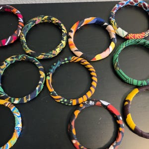 May include: A collection of fabric-covered bangles in various colors and patterns. The bracelets feature vibrant hues of red, yellow, blue, and green. The bangles are arranged on a dark surface.
