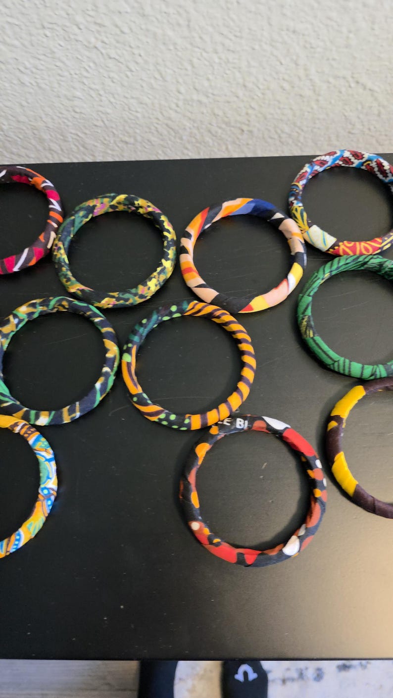 May include: A collection of fabric-covered bangles in various colors and patterns. The bracelets feature vibrant hues of red, yellow, blue, and green. The bangles are arranged on a dark surface.