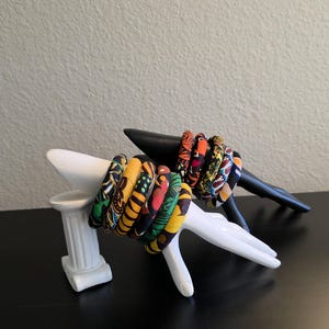 May include: A collection of colorful fabric-covered bangles displayed on a white hand-shaped sculpture and a black sculpture. The bangles feature vibrant patterns in shades of yellow, green, red, and orange. The display is set against a neutral background.
