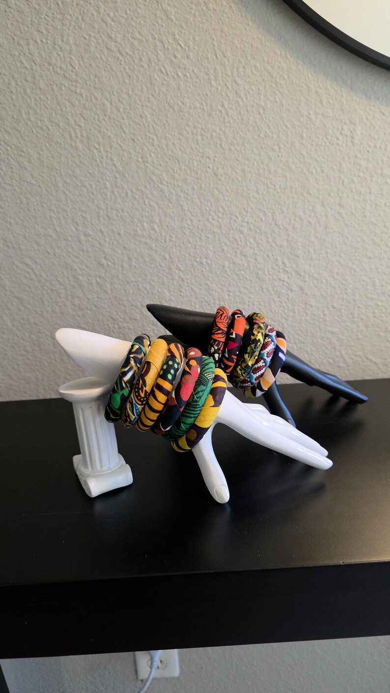May include: A collection of colorful fabric-covered bangles displayed on a white hand-shaped sculpture and a black sculpture. The bangles feature vibrant patterns in shades of yellow, green, red, and orange. The display is set against a neutral background.