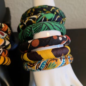 May include: A stack of fabric-covered bangles in various patterns and colors, including green, yellow, and blue. The bracelets are displayed on a white hand-shaped jewelry holder. The bangles have a tribal or African-inspired aesthetic.