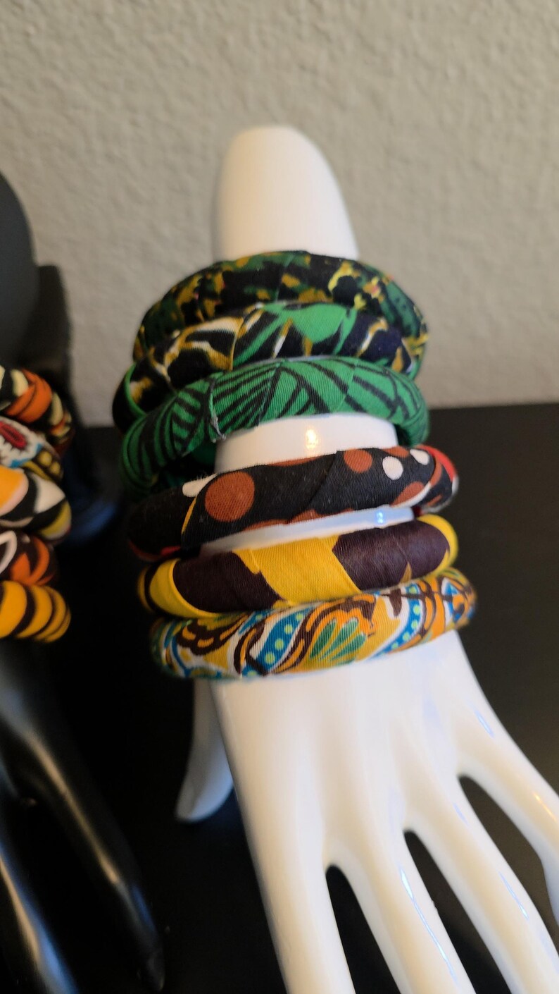 May include: A stack of fabric-covered bangles in various patterns and colors, including green, yellow, and blue. The bracelets are displayed on a white hand-shaped jewelry holder. The bangles have a tribal or African-inspired aesthetic.