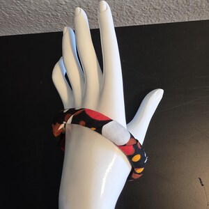 May include: A fabric-covered bracelet with a repeating pattern of red, white, yellow, and brown circles on a black background. The bracelet is displayed on a white hand-shaped mannequin.