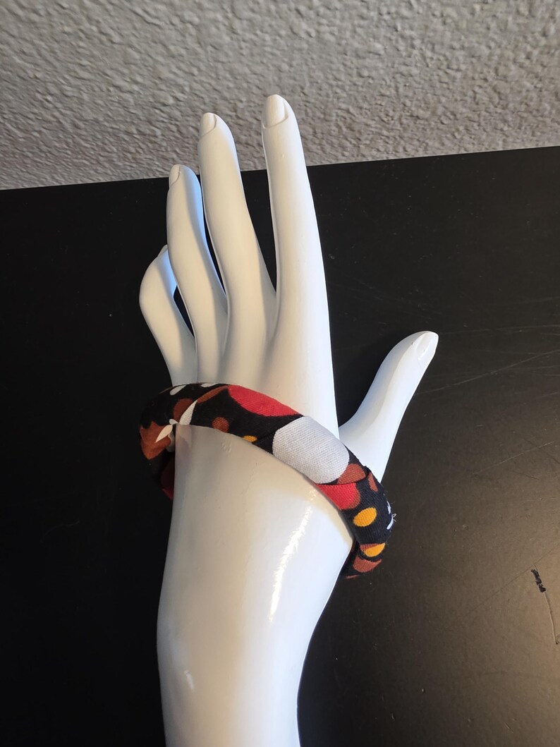 May include: A fabric-covered bracelet with a repeating pattern of red, white, yellow, and brown circles on a black background. The bracelet is displayed on a white hand-shaped mannequin.
