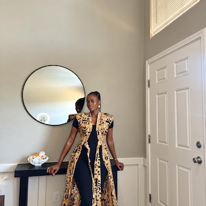 May include: A sleeveless, floor-length duster vest featuring a bold, geometric black and tan patterned print. The garment is worn open over a solid black, short-sleeved jumpsuit, creating a layered look with a structured collar and long, flowing panels.