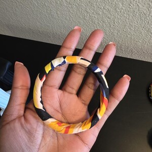 May include: A colorful fabric-covered bangle bracelet. The bracelet features a repeating pattern of yellow, black, red, and blue. The bangle is held in a hand, showcasing its design and texture. The bracelet is a fashion accessory.