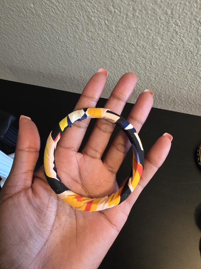 May include: A colorful fabric-covered bangle bracelet. The bracelet features a repeating pattern of yellow, black, red, and blue. The bangle is held in a hand, showcasing its design and texture. The bracelet is a fashion accessory.