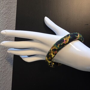 May include: A fabric-covered bangle bracelet with a colorful floral pattern in shades of black, green, yellow, and pink. The bracelet is displayed on a white mannequin hand against a black background.