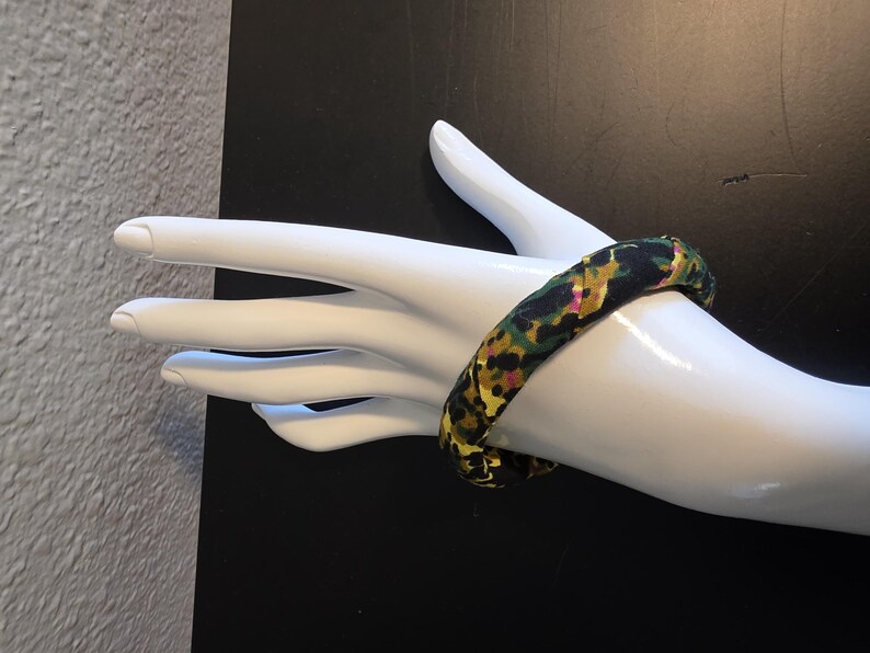 May include: A fabric-covered bangle bracelet with a colorful floral pattern in shades of black, green, yellow, and pink. The bracelet is displayed on a white mannequin hand against a black background.