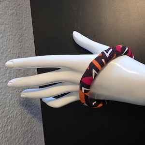 May include: A fabric-covered bangle bracelet with a geometric pattern in shades of orange, pink, white, and dark brown. The bracelet is displayed on a white mannequin hand against a black background. The bangle is a bold accessory.