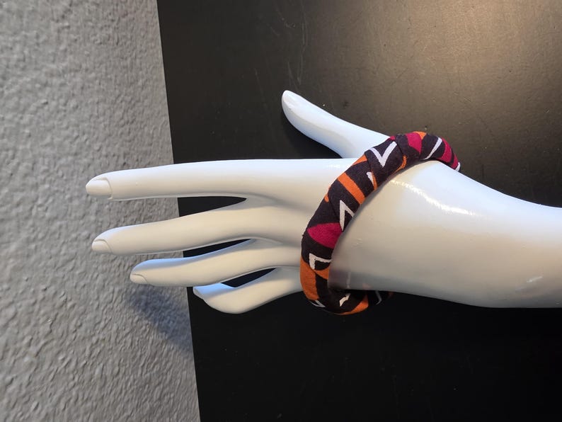 May include: A fabric-covered bangle bracelet with a geometric pattern in shades of orange, pink, white, and dark brown. The bracelet is displayed on a white mannequin hand against a black background. The bangle is a bold accessory.