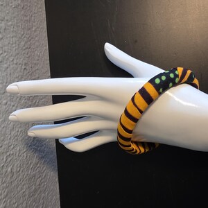 May include: A fabric-covered bracelet with a bold pattern of yellow and black stripes, accented with green polka dots. The bracelet is displayed on a white mannequin hand, showcasing its design and texture. The jewelry piece is a vibrant accessory.
