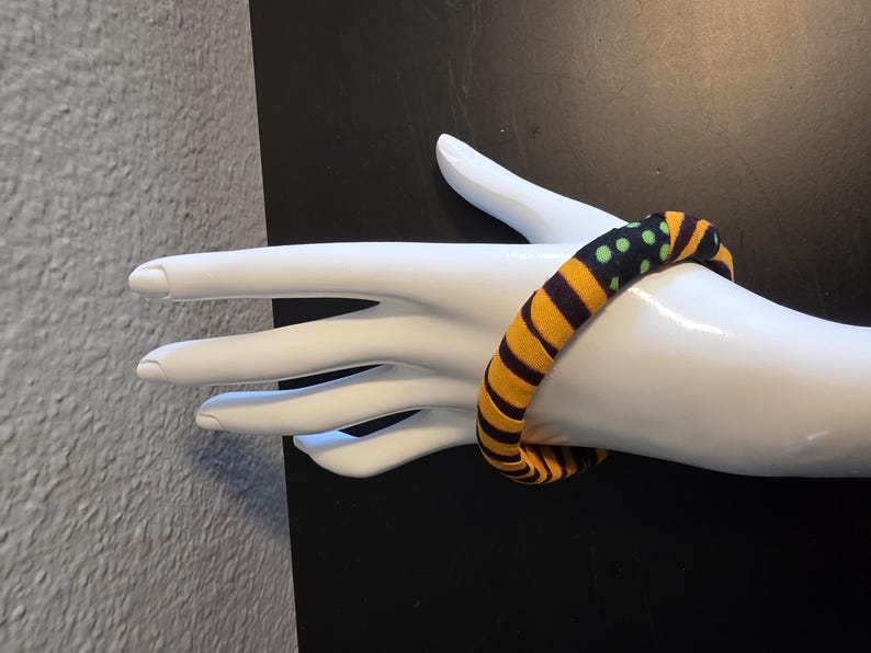 May include: A fabric-covered bracelet with a bold pattern of yellow and black stripes, accented with green polka dots. The bracelet is displayed on a white mannequin hand, showcasing its design and texture. The jewelry piece is a vibrant accessory.