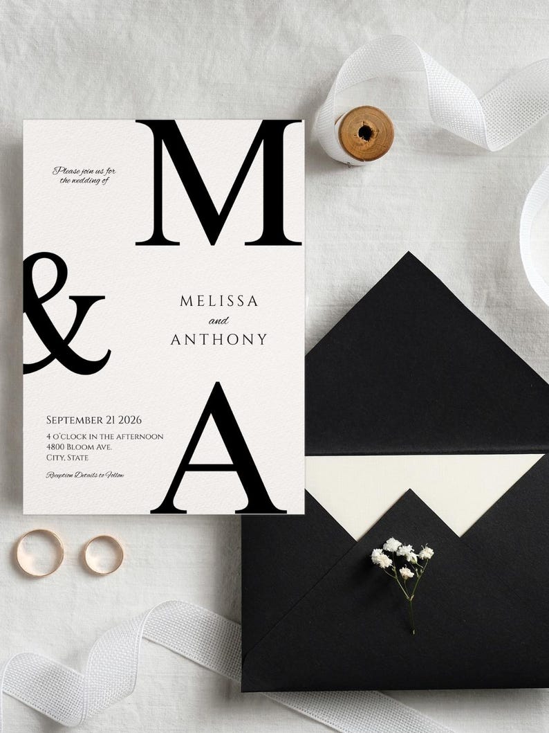 May include: Wedding invitation suite featuring a cream-colored invitation card with black text, including the names "Melissa and Anthony" and the date "September 21 2026." Includes two gold rings, a black envelope, and white ribbon.