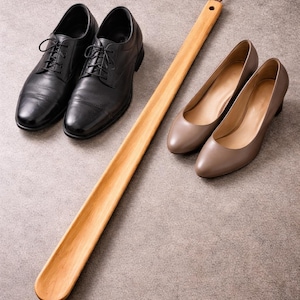 May include: A pair of black leather dress shoes with laces, a pair of taupe heels, and a long wooden shoe horn with a leather loop. The items are arranged on a gray surface.