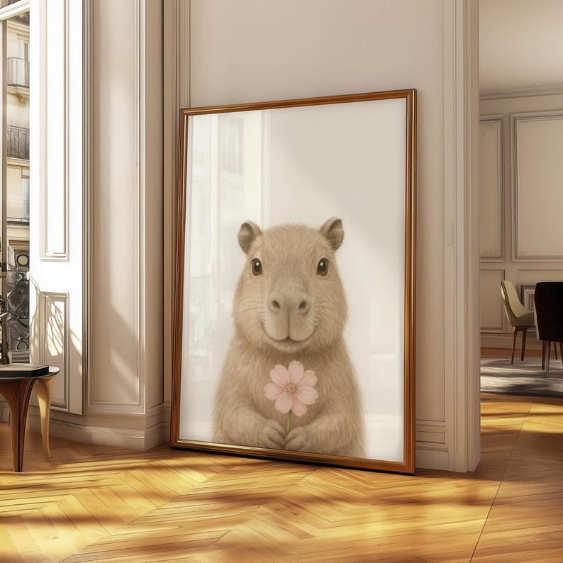 May include: A framed print of a capybara holding a pink flower. The capybara has light brown fur and a gentle expression. The artwork is set in a gold-coloured frame, adding a touch of elegance to the piece. The print is displayed in a room with wooden flooring.