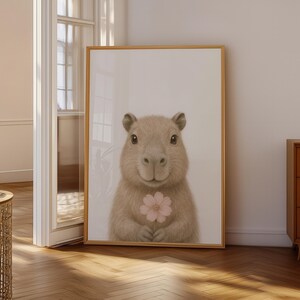 May include: A framed print of a capybara holding a pink flower. The capybara has soft, brown fur and a gentle expression. The artwork is set against a neutral background, framed in a light wood, suitable for a child's room.