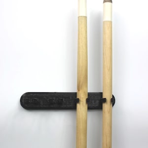 May include: A black wall-mounted cue rack holding two wooden cues. The cues have white tips and brown ends. The rack is mounted on a white wall.